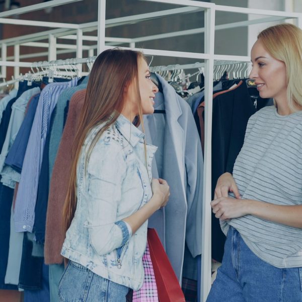 Two women chatting among clothing racks in a stylish fashion store, sharing shopping advice.